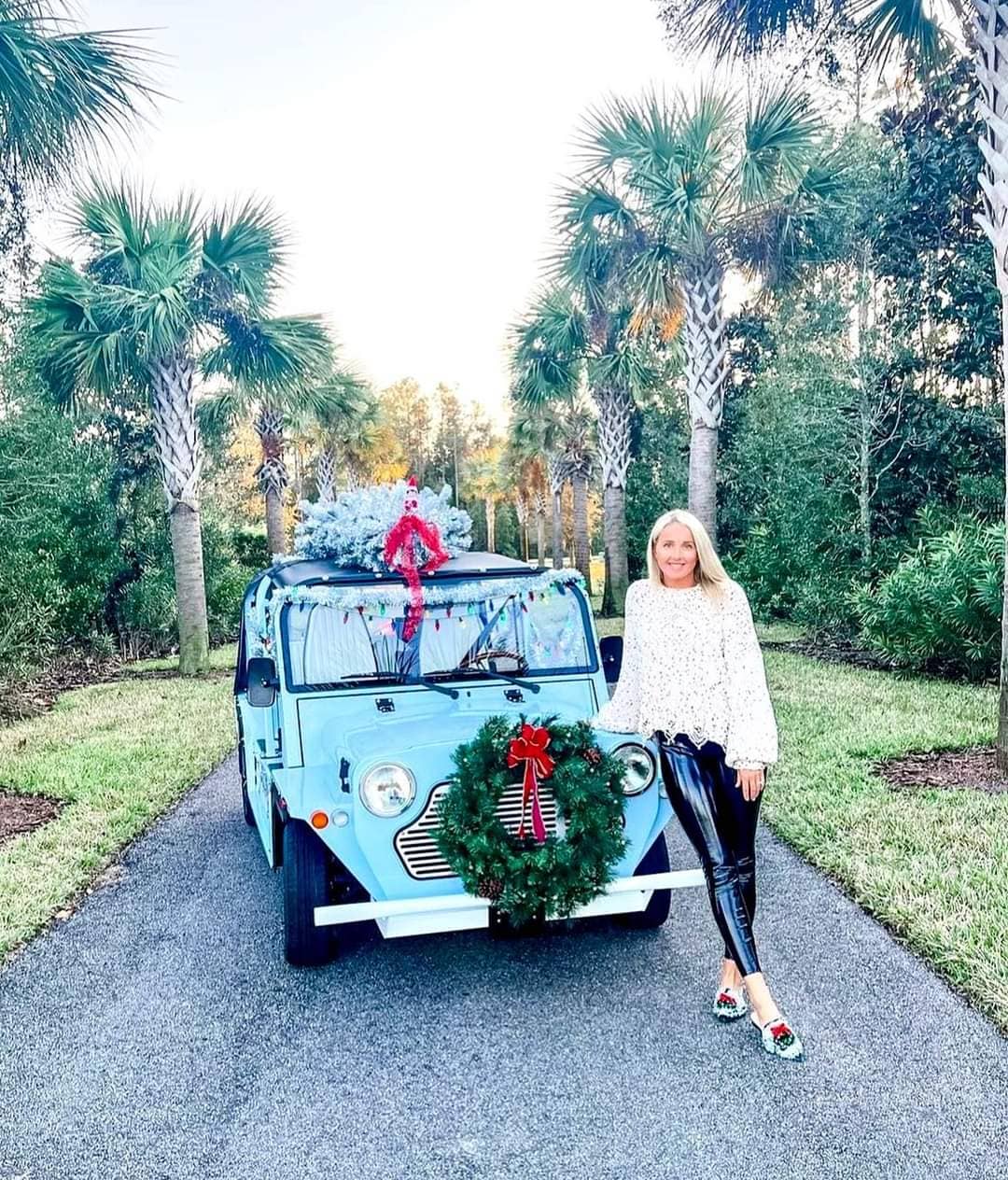 A woman stands beside a light blue car decorated with a wreath and gifts, surrounded by palm trees on a sunny day.