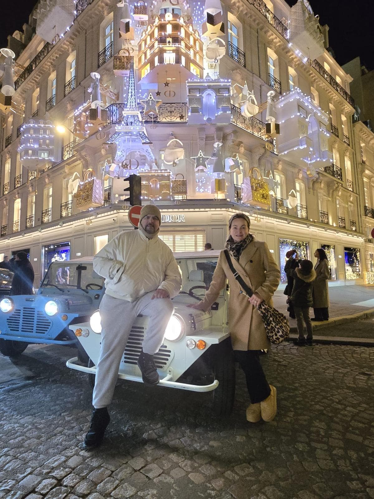 Two people pose by a white car on a cobblestone street, with a building decorated with festive lights in the background.