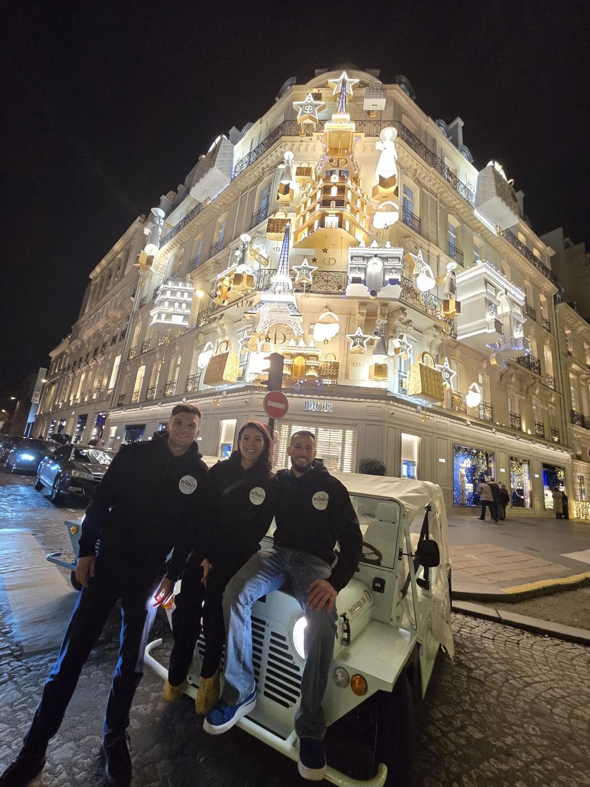 Three people pose in front of a brightly lit, elaborately decorated building at night, with festive lights and ornaments with Mini Moke.