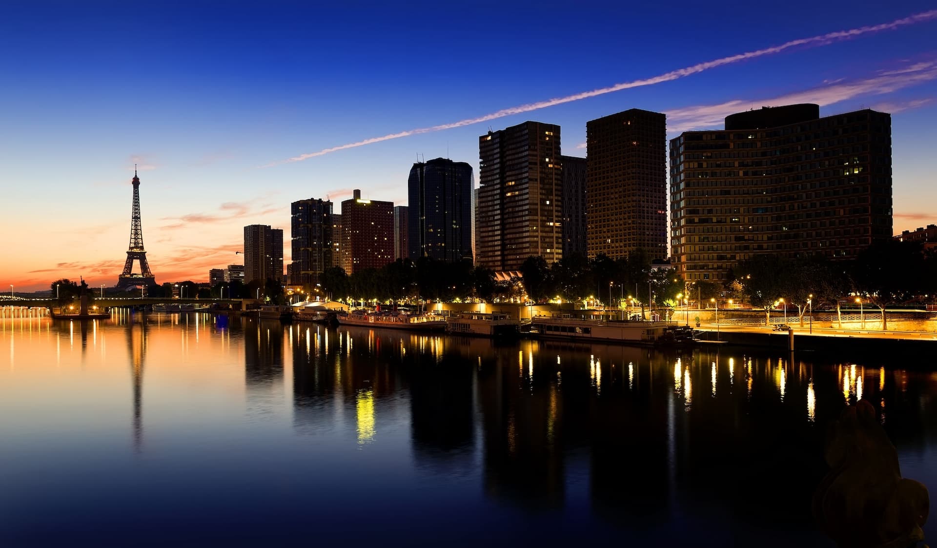 Paris skyline at dusk with the Eiffel Tower and illuminated buildings reflected in the Seine River under a twilight sky.