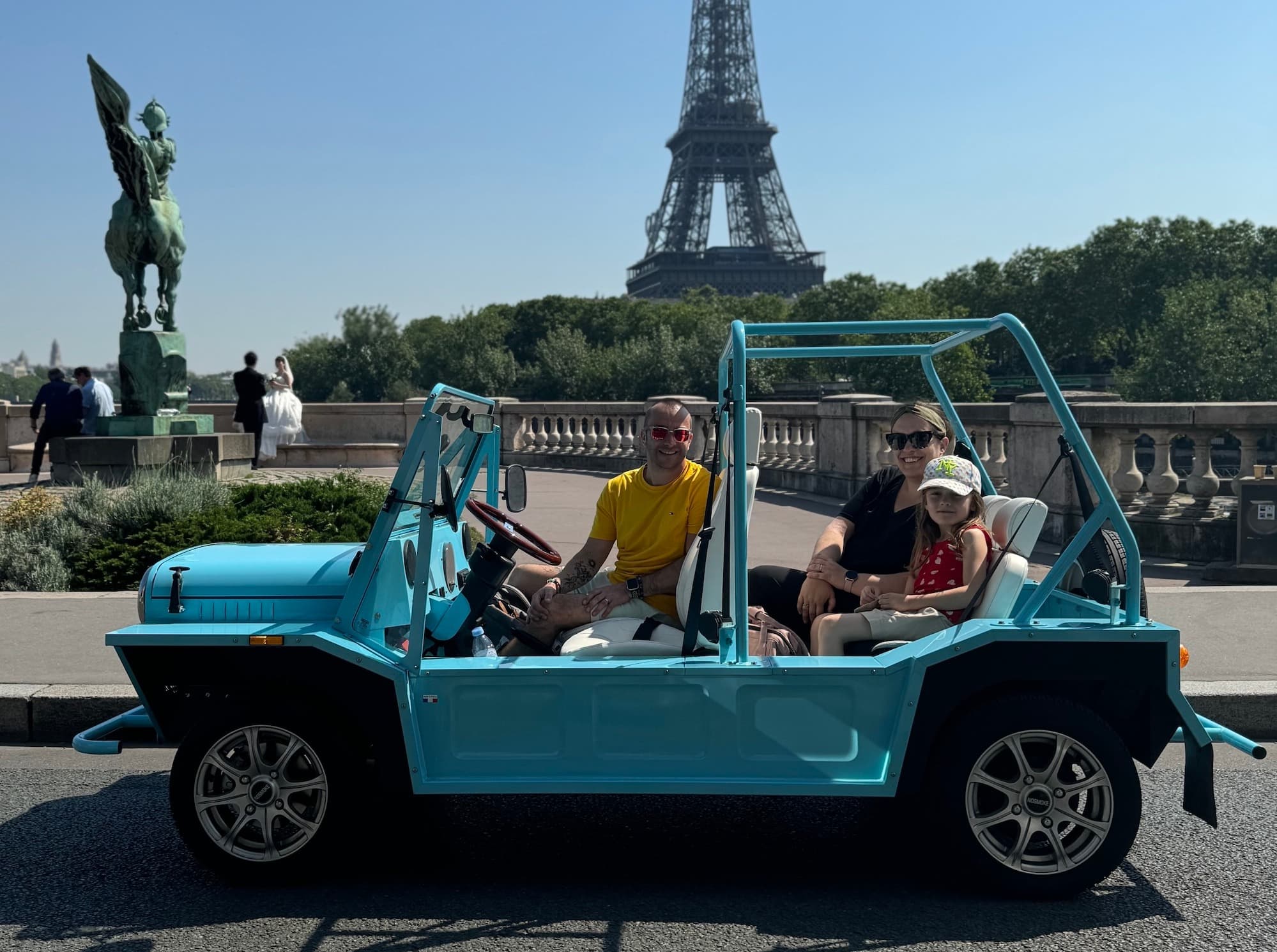 A family in a blue open-top Mini Moke poses near the Eiffel Tower, with a statue and trees in the background.