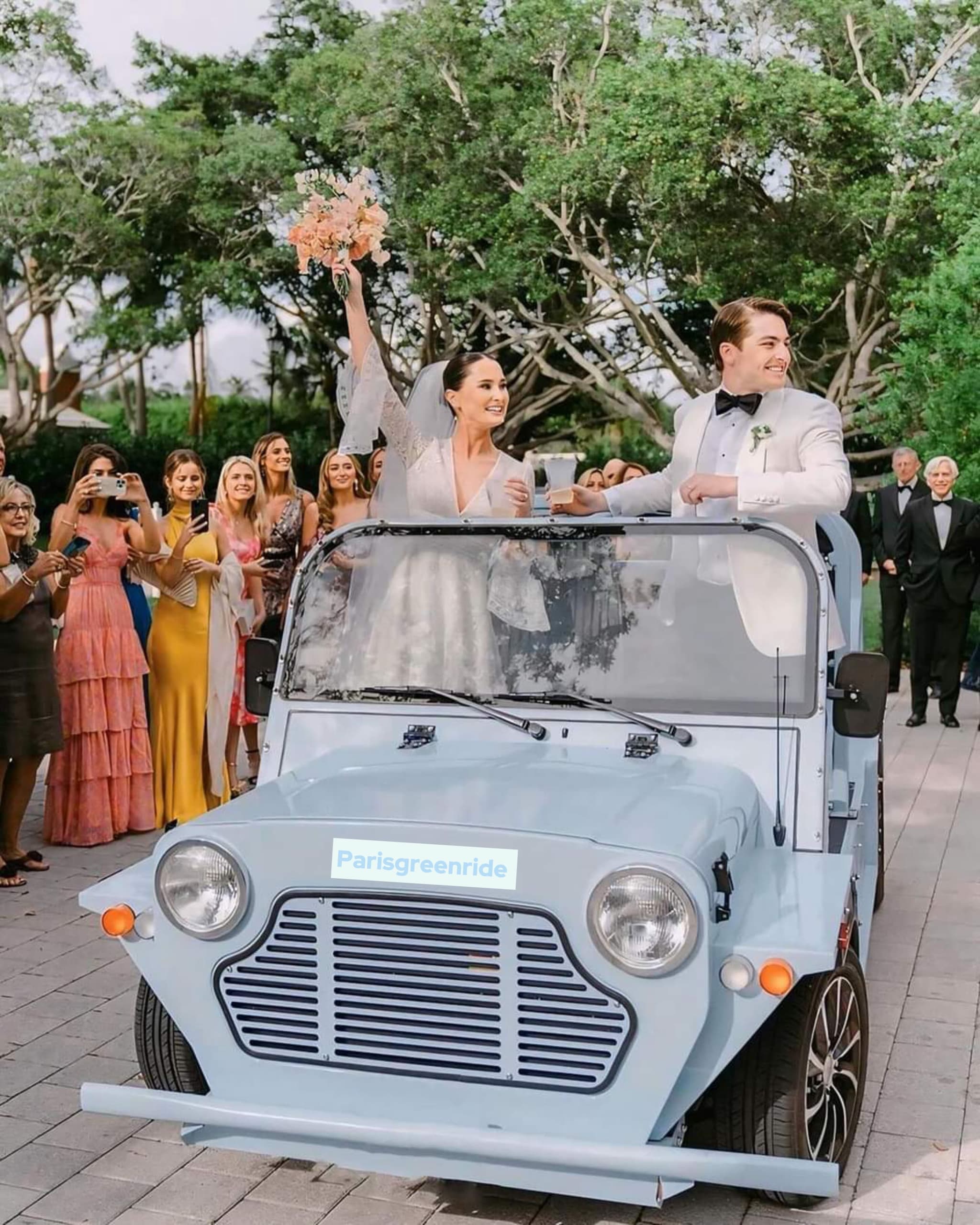 Bride and groom joyfully ride in a Mini Moke small blue car, surrounded by cheering guests with trees in the background.