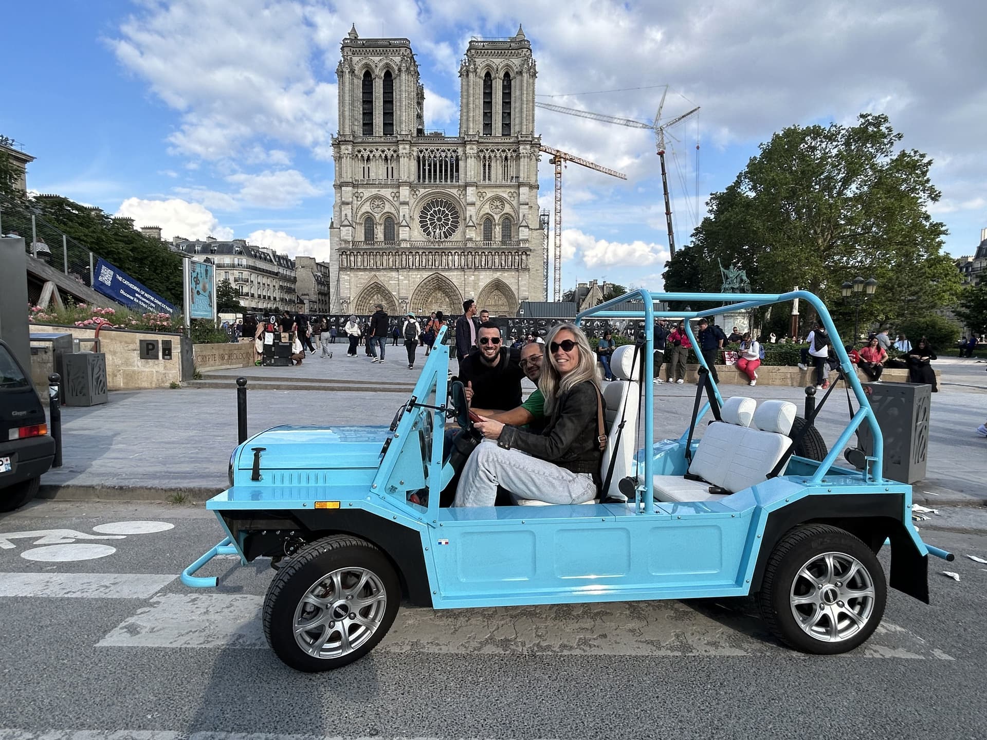 Three people in a blue open-top Mini Moke in front of Notre-Dame Cathedral, Paris, on a sunny day.