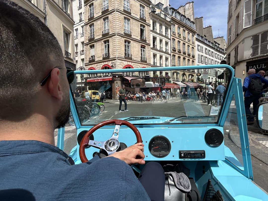 A person drives a vintage blue mini moke through a bustling European street with historic buildings and outdoor cafes visible ahead.