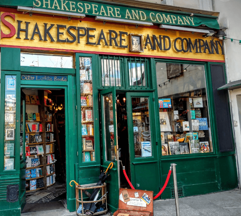 Front view of Shakespeare and Company bookstore in Paris, with green facade, books displayed in windows, and a small outdoor book cart.