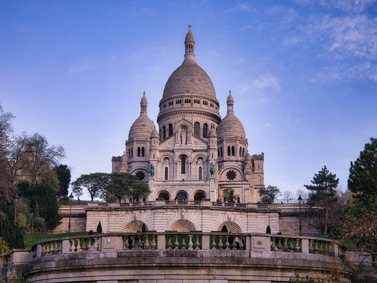 The Basilica of Sacré-Cœur with its domes and intricate architecture, set against a clear blue sky, surrounded by greenery.
